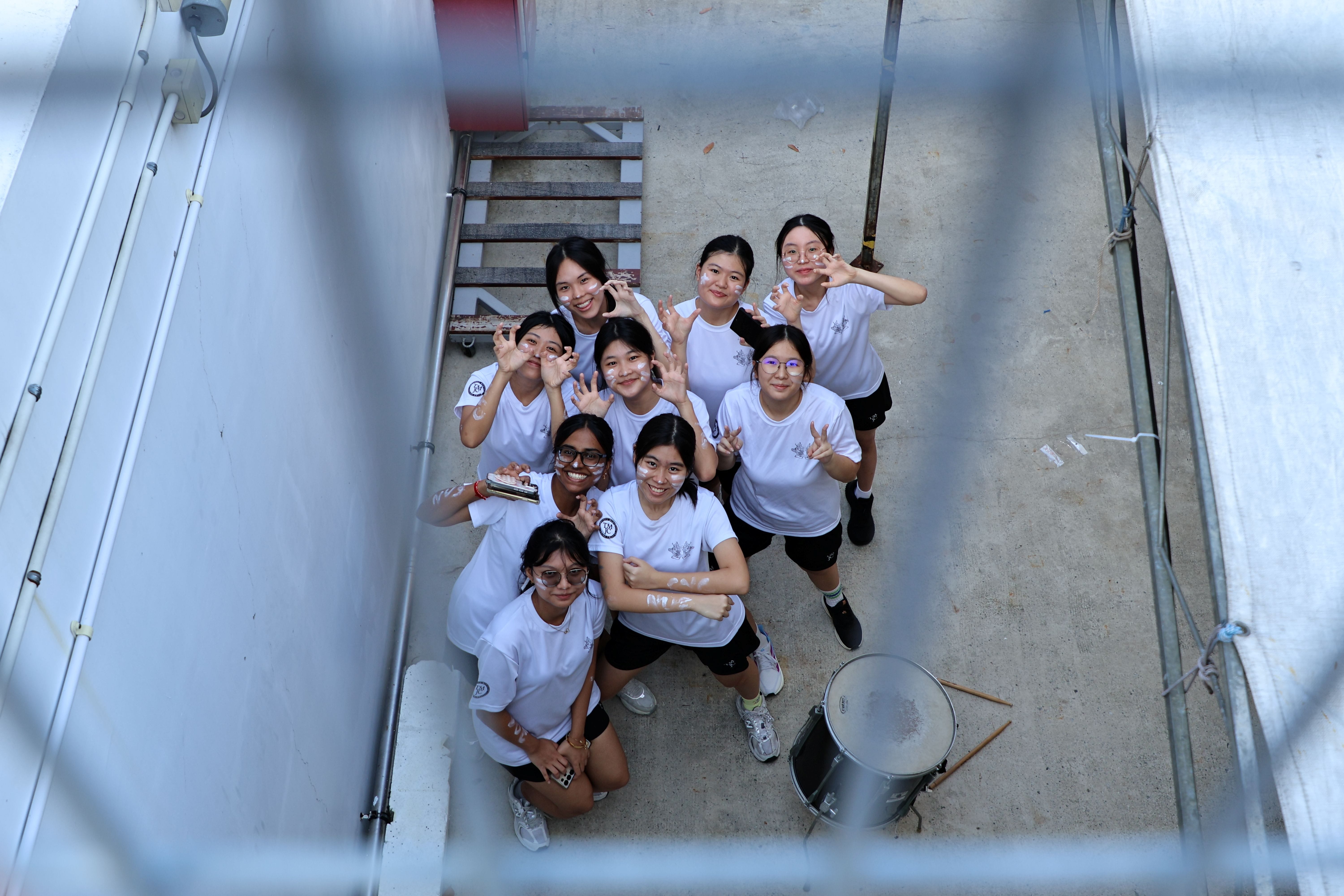 Nine female students in white T-shirts with face paint look up at the camera from a stairwell, waving and smiling, with a snare drum beside them.
